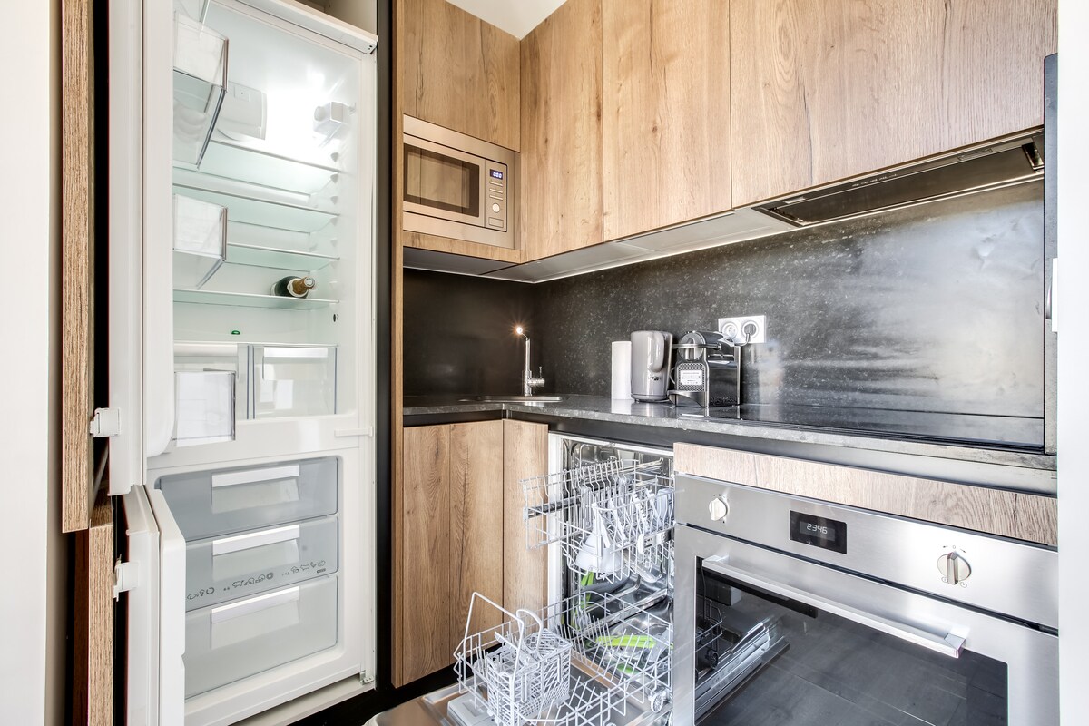 A fully equipped kitchen features modern appliances, including a refrigerator, microwave, and oven. A dishwasher is visible in the lower cabinet, while a countertop provides workspace. The cabinetry showcases a warm wood finish, complementing the sleek black backsplash.