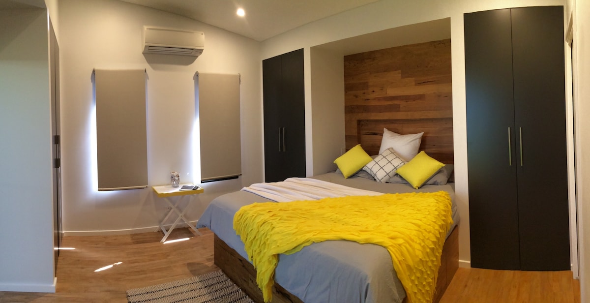 A serene bedroom showcases a spacious bed adorned with bright yellow and gray linens. The room features a wooden accent wall and built-in wardrobes. Natural light filters through two window shades, enhancing the calm ambiance, while light-colored flooring adds to the overall inviting look.