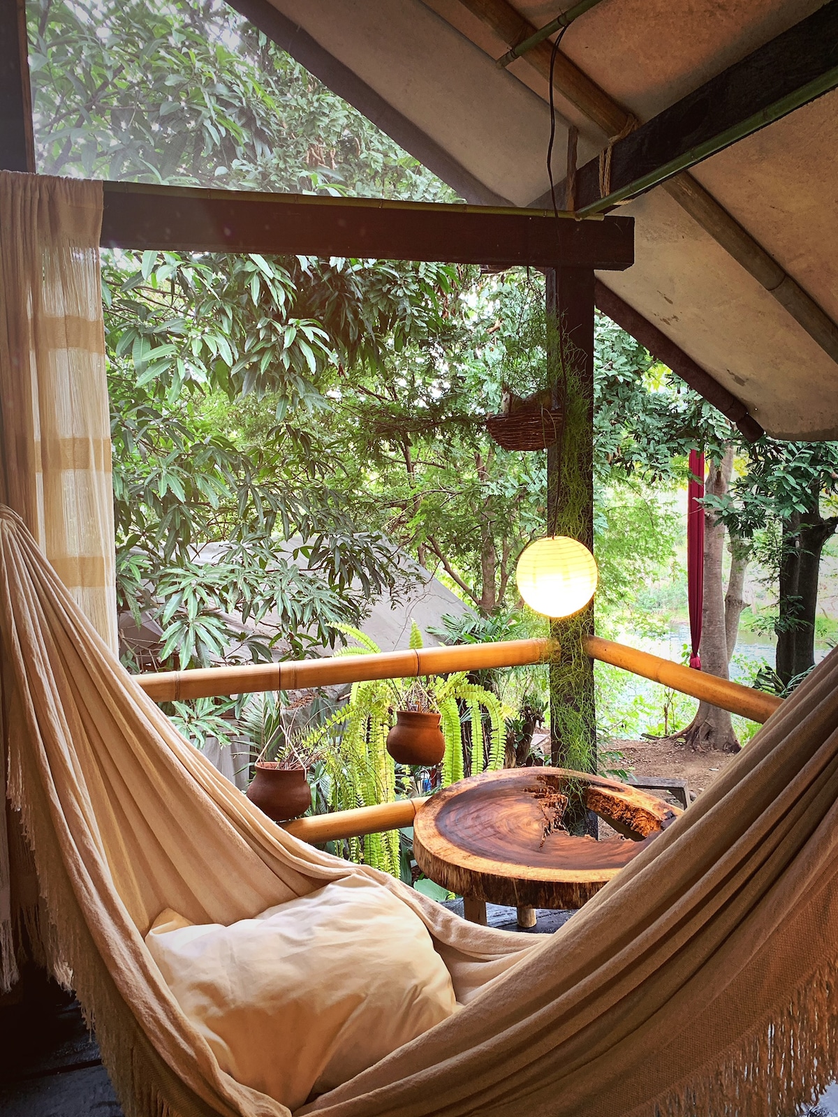 A hammock is draped on a wooden balcony, inviting relaxation among the lush greenery. A natural wood table complements the outdoor space, while a round lantern provides soft lighting. Surrounding foliage enhances the tranquil setting, offering views of the vibrant landscape.