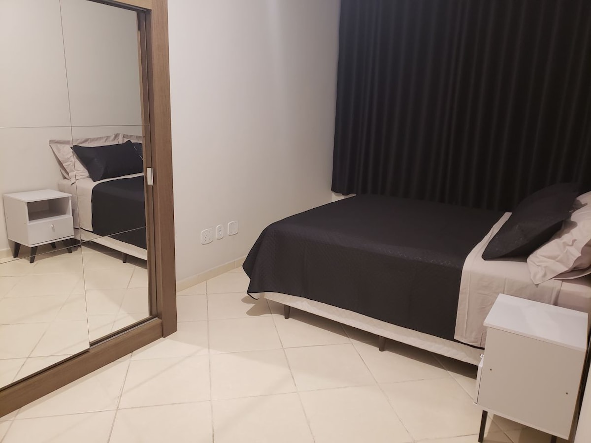 A serene bedroom space features a spacious bed dressed in dark bedding, complemented by a bedside table. A large mirror reflects the room’s modern design, while dark curtains drape over the window, allowing control of natural light.