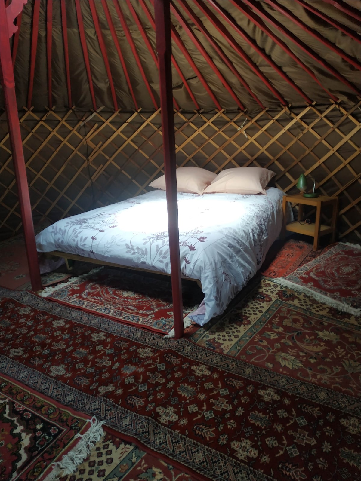 A spacious bed is positioned in the center of the yurt, featuring light-colored bedding with decorative patterns. Soft pillows rest atop the bed, and a small wooden table is placed nearby. Richly colored rugs cover the floor, enhancing the natural ambiance.