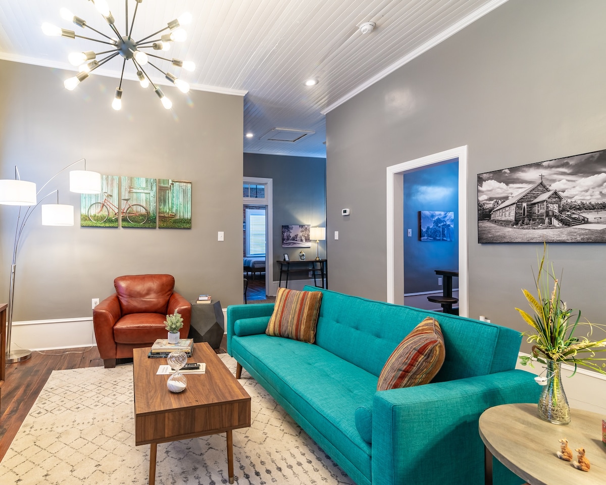 A colorful teal sofa is placed in the living area, accompanied by a brown leather chair. A wooden coffee table sits in front, adorned with a small plant. The walls are painted in soft gray, and the space is enhanced by artistic wall decor and modern lighting.