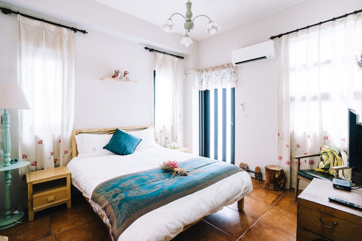 A spacious bedroom features a large bed adorned with a decorative blue accent pillow and a flowery throw. Natural light filters through sheer curtains on multiple windows. A bedside lamp and wooden drawer provide functional decor, while a small television and wooden furniture complete the inviting atmosphere.