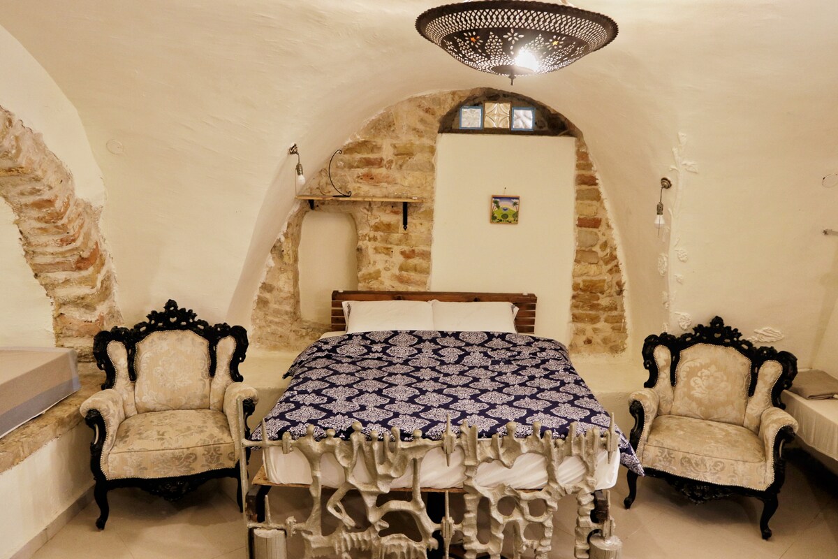 A spacious room features a large, patterned bedspread covering the bed. Two ornate armchairs with dark wood frames are positioned near the bed. The walls, displaying exposed stone accents, are complemented by a decorative ceiling light fixture, creating a warm and welcoming setting.