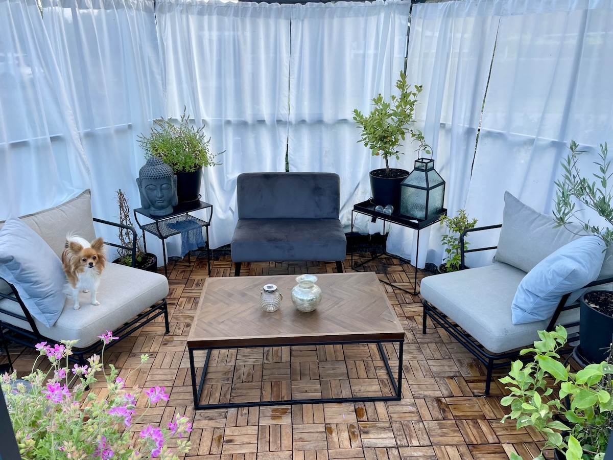 An inviting seating area is enclosed by sheer white curtains, featuring a small coffee table and two armchairs with soft blue cushions. A gray sofa complements the setting, surrounded by potted plants that add greenery to the cozy space.