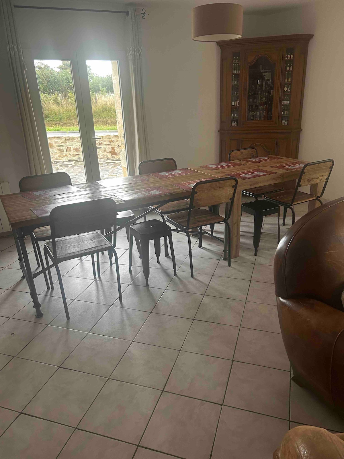 A spacious dining area is featured, highlighted by a long, wooden table surrounded by several metal chairs. Light enters through large glass doors that open to a natural landscape, creating a relaxed atmosphere. A wooden cabinet is positioned against the wall, adding functional storage.