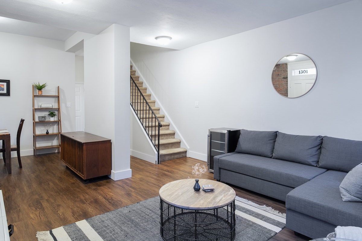 The living area features a gray sectional sofa, complemented by a round coffee table on a patterned rug. A mirror adorns the wall, reflecting natural light. In the background, a dining area with a wooden table and a staircase leads to the upper floor.