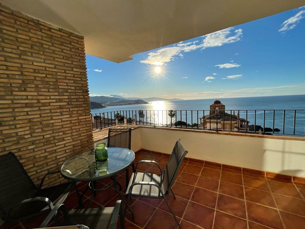 A terrace offers an unobstructed view of the ocean, illuminated by the soft glow of the setting sun. A glass table with metal chairs is positioned on a tiled surface, while a low wall adorned with wrought iron details frames the scene.