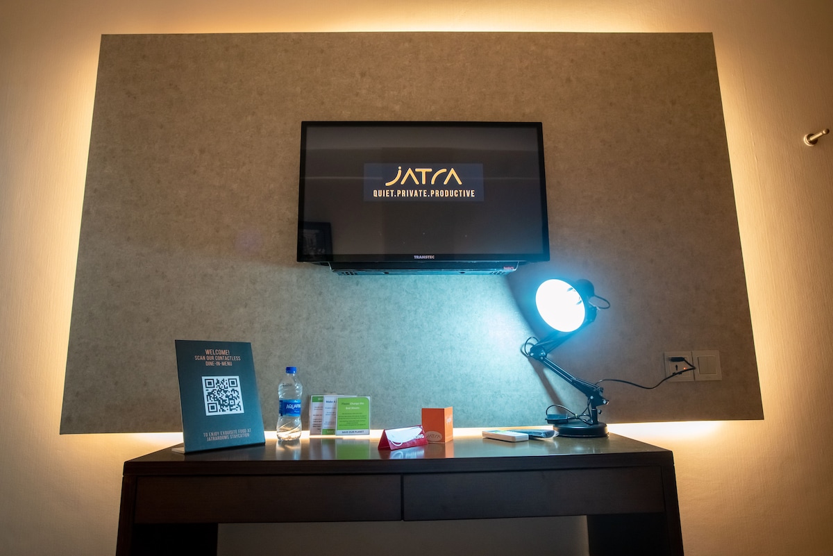 A wall-mounted LED TV is positioned above a sleek wooden desk, accompanied by a task lamp. A bottle of water and assorted stationery are placed on the desk. Soft ambient lighting highlights the modern design of the area.
