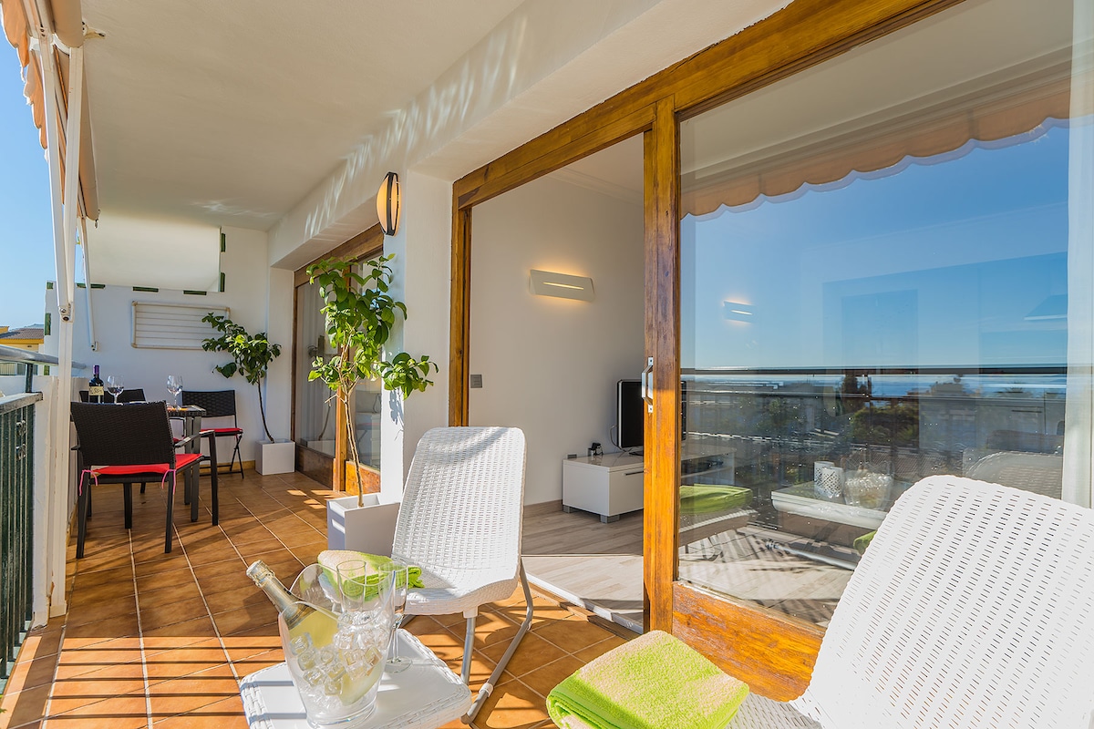 The terrace features comfortable lounge chairs, overlooking a vibrant sea view. Natural light floods the indoor area through expansive glass doors, which offer a seamless transition between the outdoor and indoor living spaces. Potted plants add a touch of greenery.