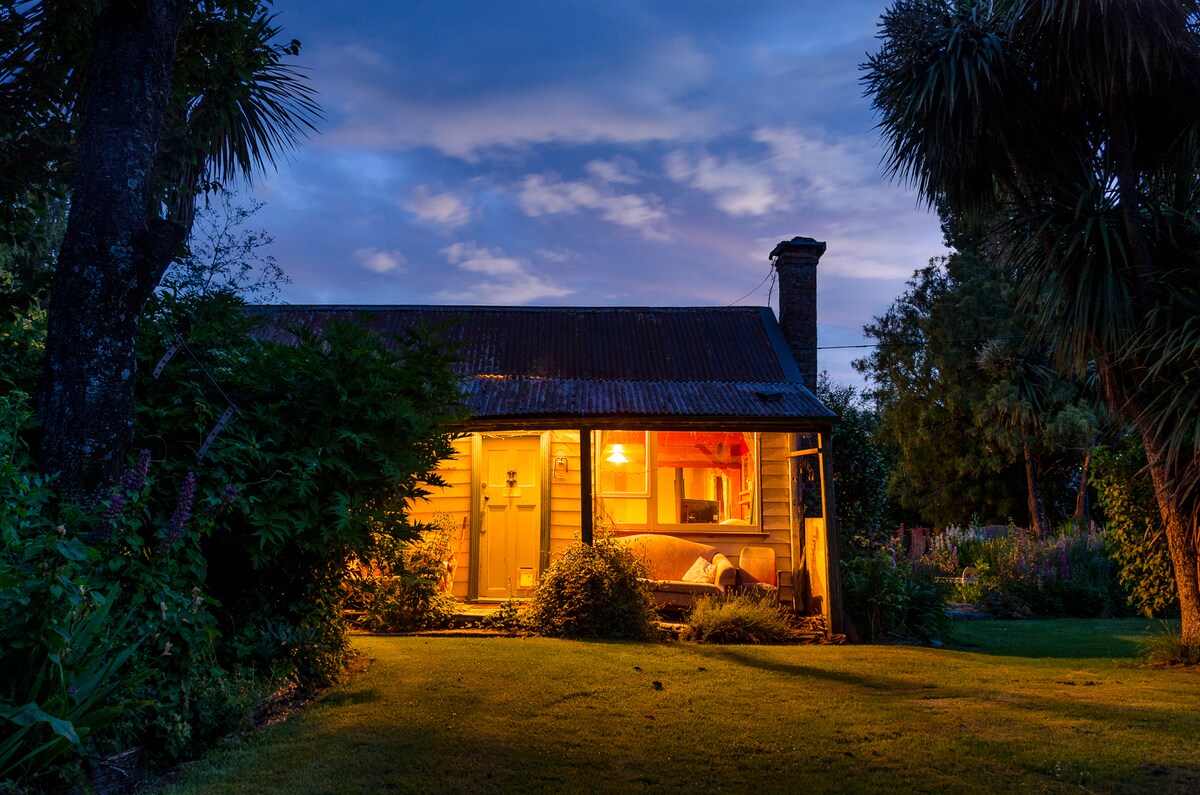 The cottage, illuminated by warm light, is framed by lush greenery and palm trees. A rustic facade with a corrugated roof is highlighted against a twilight sky, creating a welcoming view of a cozy retreat. The surrounding garden adds to the serene atmosphere.