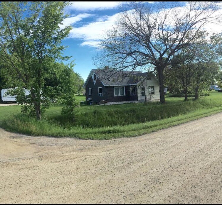 A charming house is set back from a gravel road, surrounded by green grass and trees. The exterior features a combination of light and dark siding, with windows allowing natural light. An inviting front porch is visible, offering a shaded spot for relaxation.