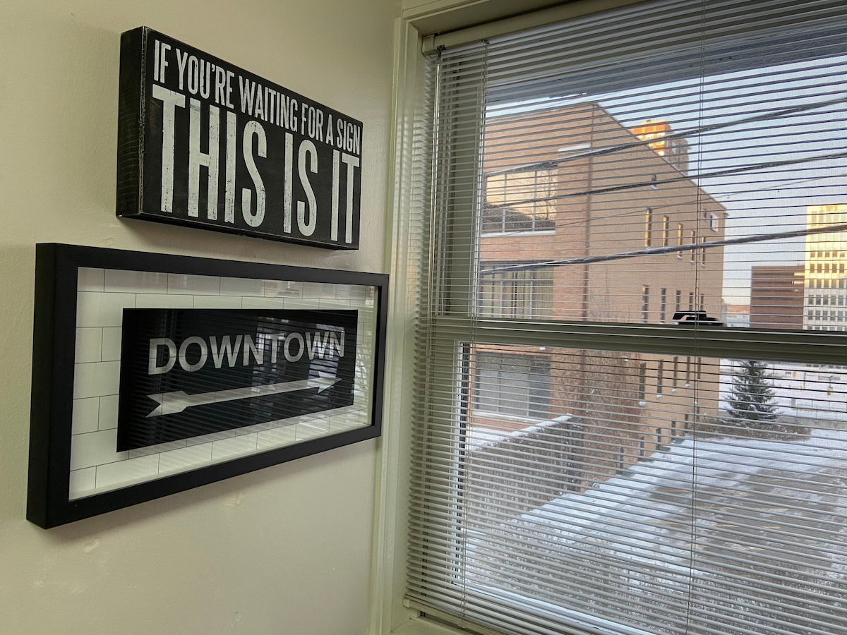 A corner of the living space features two framed signs, one stating 'THIS IS IT' and the other reading 'DOWNTOWN.' Natural light filters through the window, illuminating the area and providing a view of the neighboring buildings.