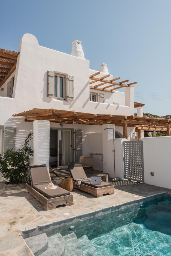 The exterior of the villa features a wooden pergola providing shade over the outdoor seating area. Sun loungers are positioned beside a small pool, while a stone pathway leads to the entrance. The light-colored walls create a contrast against the blue sky.