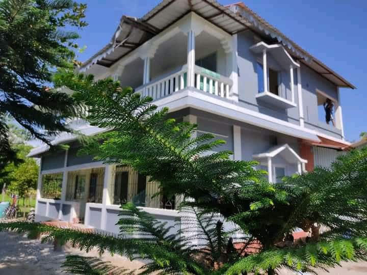 A two-story building with a light gray exterior features a balcony on the second level and a wraparound porch on the ground floor. Lush greenery partially frames the house, enhancing its inviting appearance under a clear blue sky.