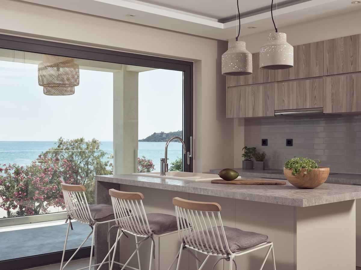 A modern kitchen features a large island with seating for four. Stylish light fixtures hang above, while large glass doors open to a scenic view of the beach and greenery. Natural light fills the space, enhancing the sleek cabinetry and minimalist design.