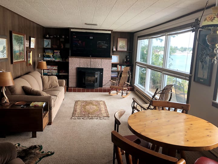 The main living area: TV above fireplace, dining table, pellet stove, and a wall of windows looking out over Lake Superior.