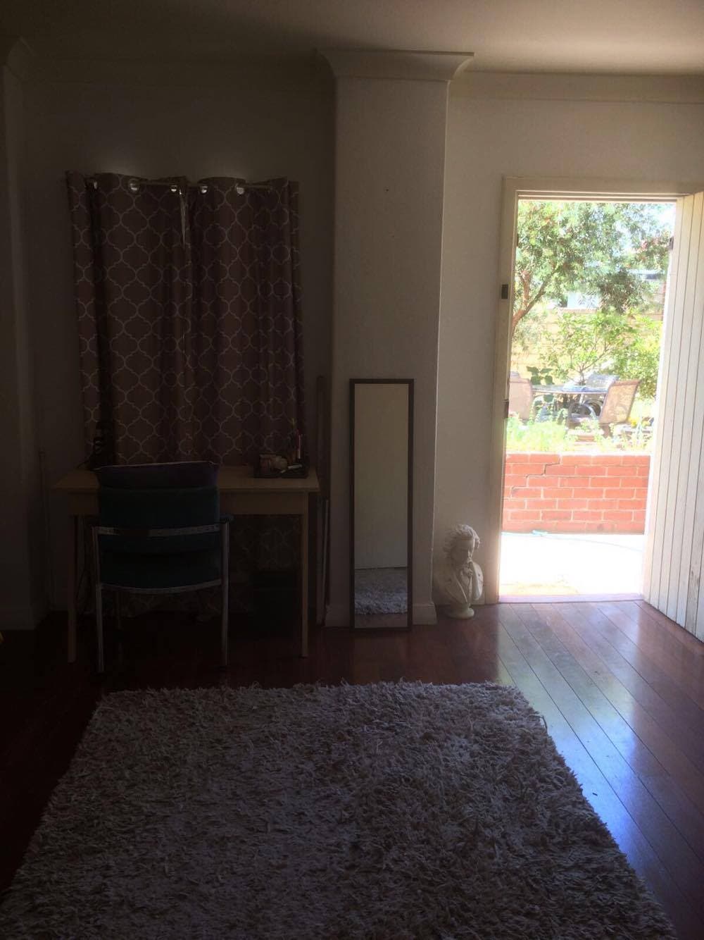 A corner of the studio features a wooden desk and a chair next to a full-length mirror. Natural light enters through a partially open door leading to a patio area. Soft beige carpet covers the floor, adding warmth to the space.