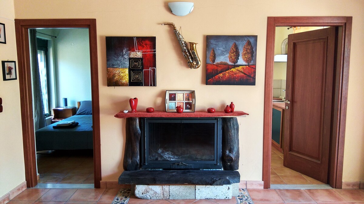 A welcoming living space is showcased, featuring a wooden fireplace with a stone base. Walls are adorned with vibrant artwork, and two doorways lead to distinct rooms. A cozy setting is enhanced by a subtle arrangement of decorative items on the mantel.