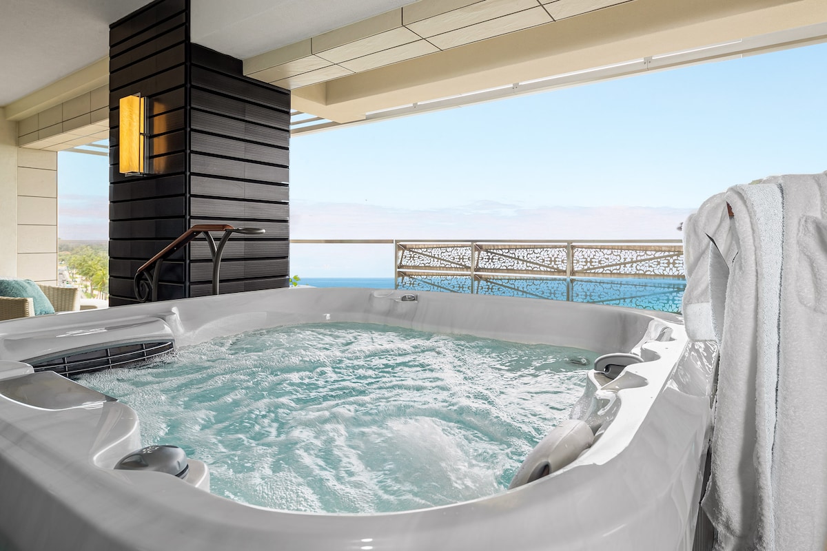 A private hot tub is set on an outdoor balcony, surrounded by a sleek railing. The bubbling water reflects the bright blue sky and provides an expansive view of the ocean, creating a serene atmosphere for relaxation.