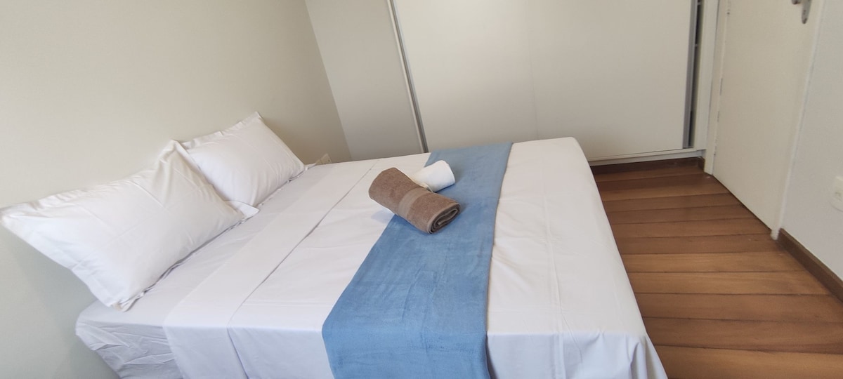 A comfortable sleeping area features a spacious bed with fresh white linens and an accent blanket in a soft blue hue. Two neatly arranged pillows and a rolled towel are placed on the bed, complementing the light wooden flooring and bright, neutral walls.
