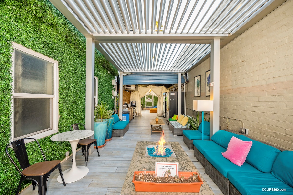 An outdoor common area features comfortable seating on a modern sofa, with bright cushions and an outdoor fire pit at the center. Greenery decorates the walls, and a round table with chairs is positioned against the vibrant backdrop, creating a relaxed atmosphere.