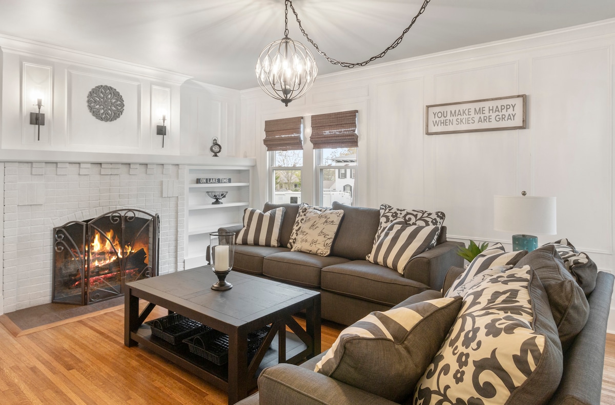 A modern living area is showcased, featuring a large sectional sofa adorned with patterned pillows. A coffee table sits at the center, while a fireplace with a white brick surround is situated against the wall. Light emanates from an elegant chandelier above, complementing the warm wooden floor.