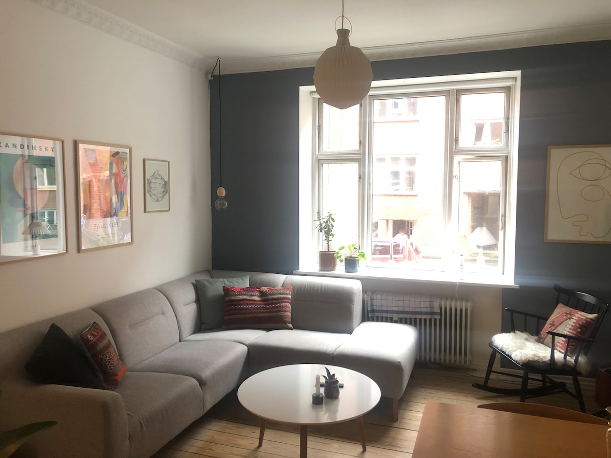 A bright living room is showcased with a cozy gray sectional sofa adorned with colorful cushions. Natural light streams in through a large window, illuminating the space. A round coffee table with a small plant sits in the center, complemented by framed art on the walls.