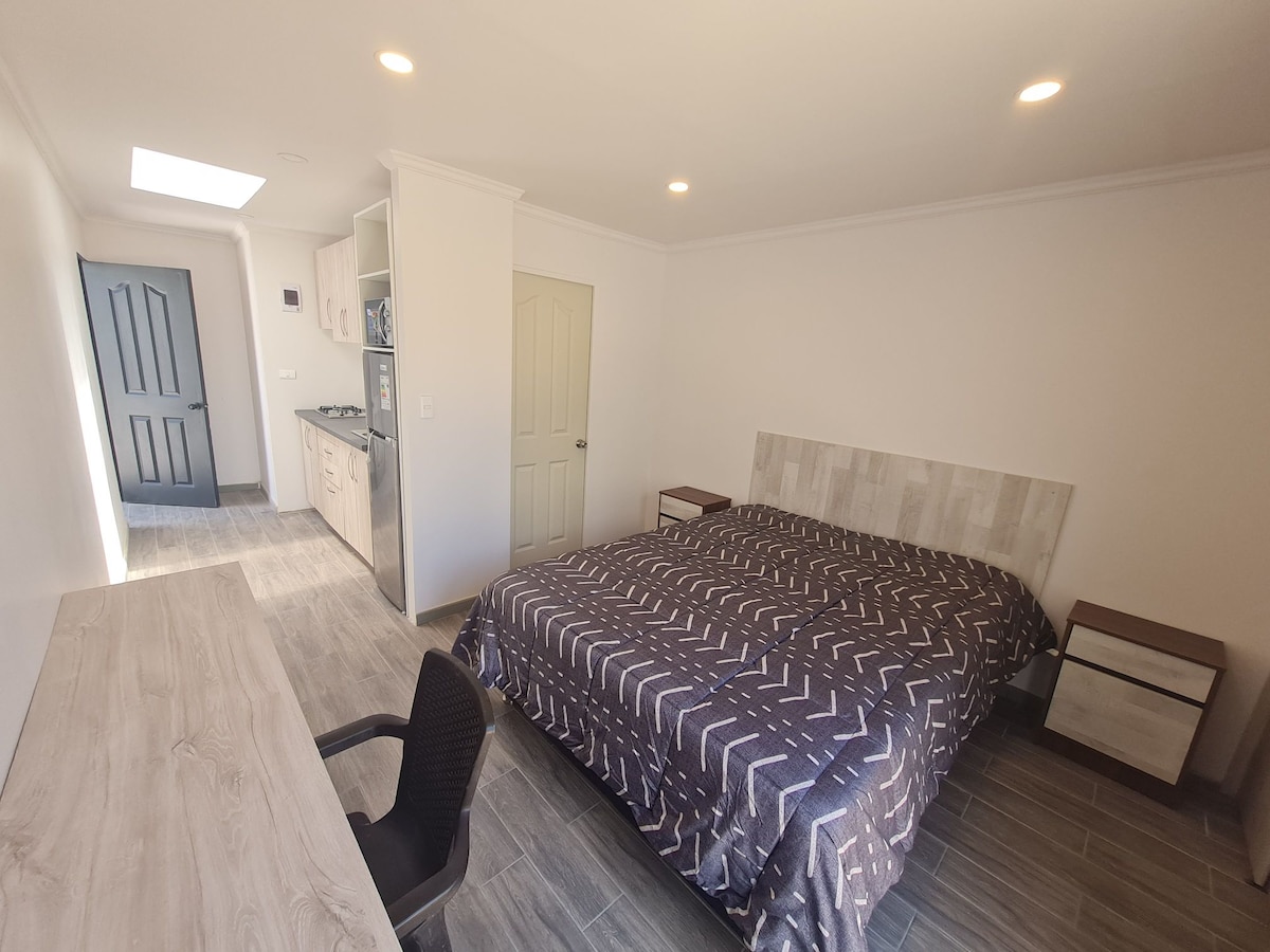 A spacious loft features a large bed covered with a patterned quilt, creating a relaxed sleeping area. A desk and chair are positioned nearby, and light enters through a skylight, illuminating the neutral-toned walls and tiled flooring. Kitchenette amenities are visible in the background.