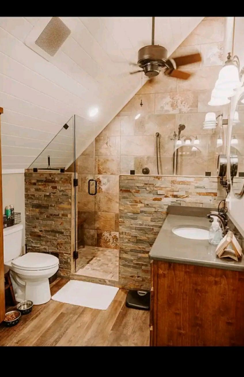A modern bathroom is displayed, featuring a spacious glass-enclosed shower with a stone accent wall. Two sinks are positioned atop a wooden vanity, complemented by a decorative mirror. Natural light filters in, enhancing the warm tones of the fixtures and flooring.