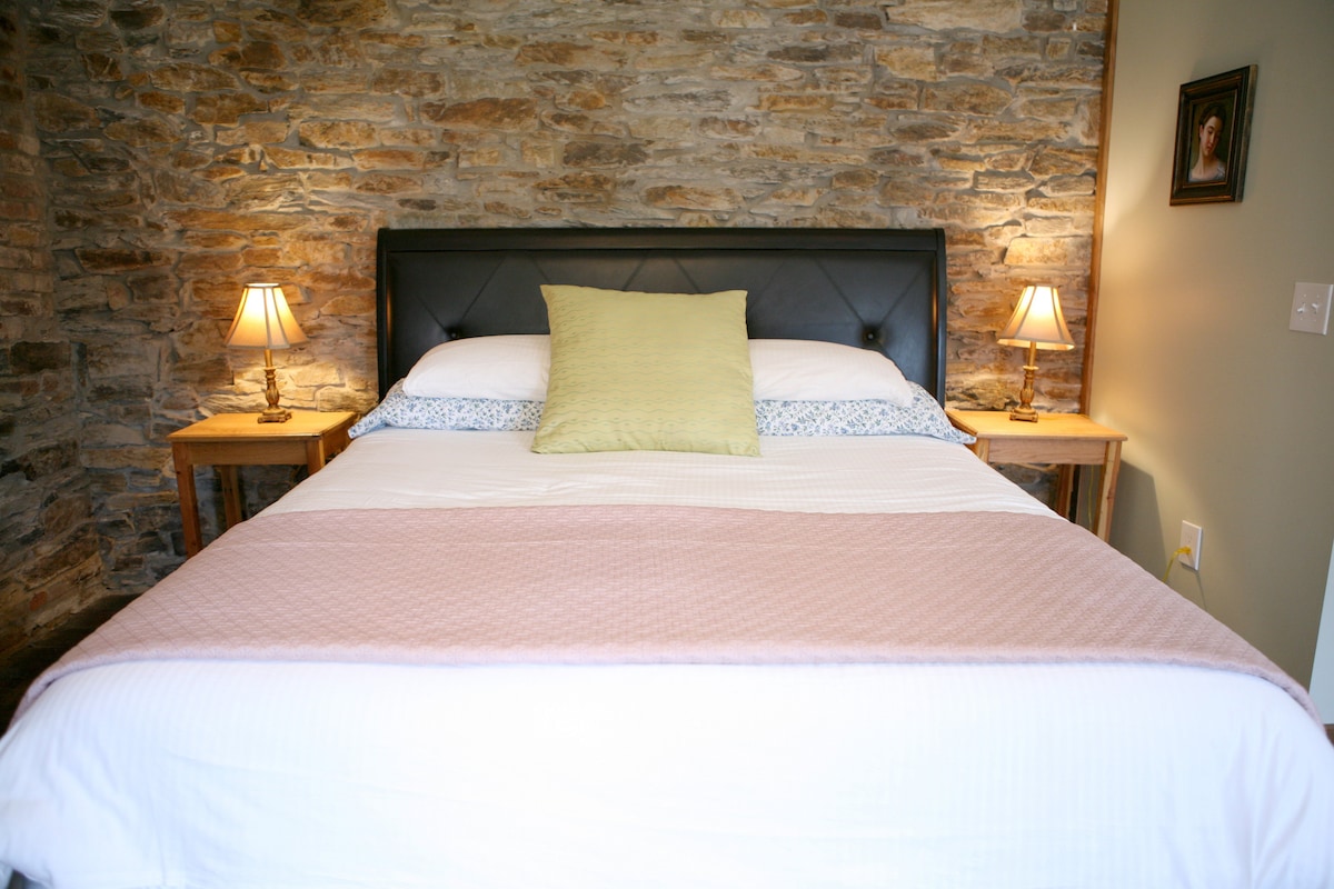 A comfortable sleeping area features a king size bed with a light pink throw and patterned accent pillows. Soft lighting is provided by two bedside lamps on wooden tables, set against rustic stone walls that reflect the room's historical charm.