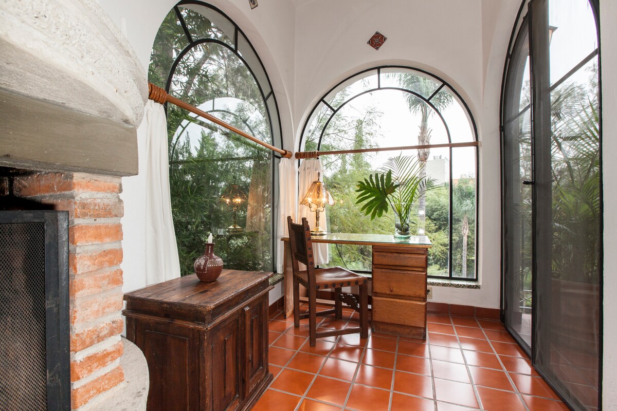 A cozy nook featuring arched windows that showcase lush greenery outside. A wooden desk and chair are positioned beside the fireplace, with a decorative vase sitting on a storage chest. Soft light filters through the curtains, enhancing the tranquil atmosphere.