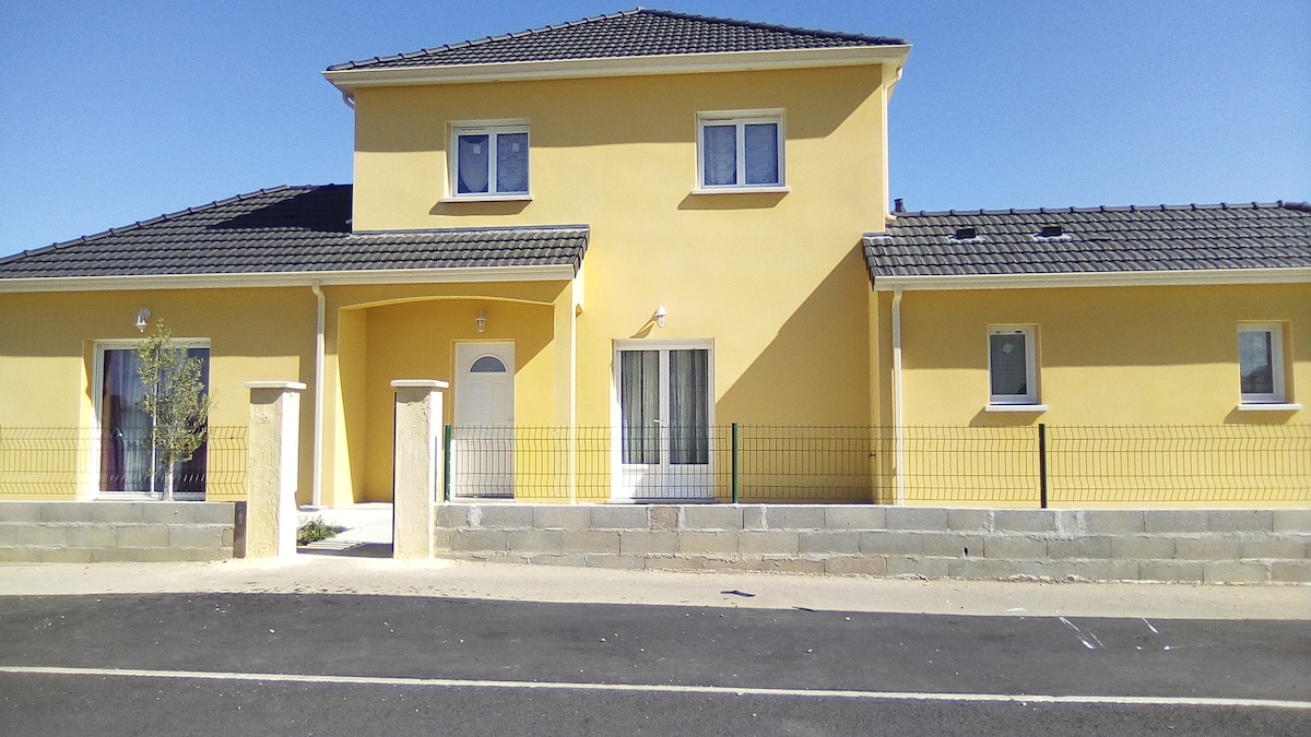 A two-story yellow house with a symmetrical facade is presented. Windows are evenly spaced along the front, with a main entrance featuring a covered porch. A low stone wall surrounds the property, enhancing the outdoor space. Clear blue skies create a bright backdrop.
