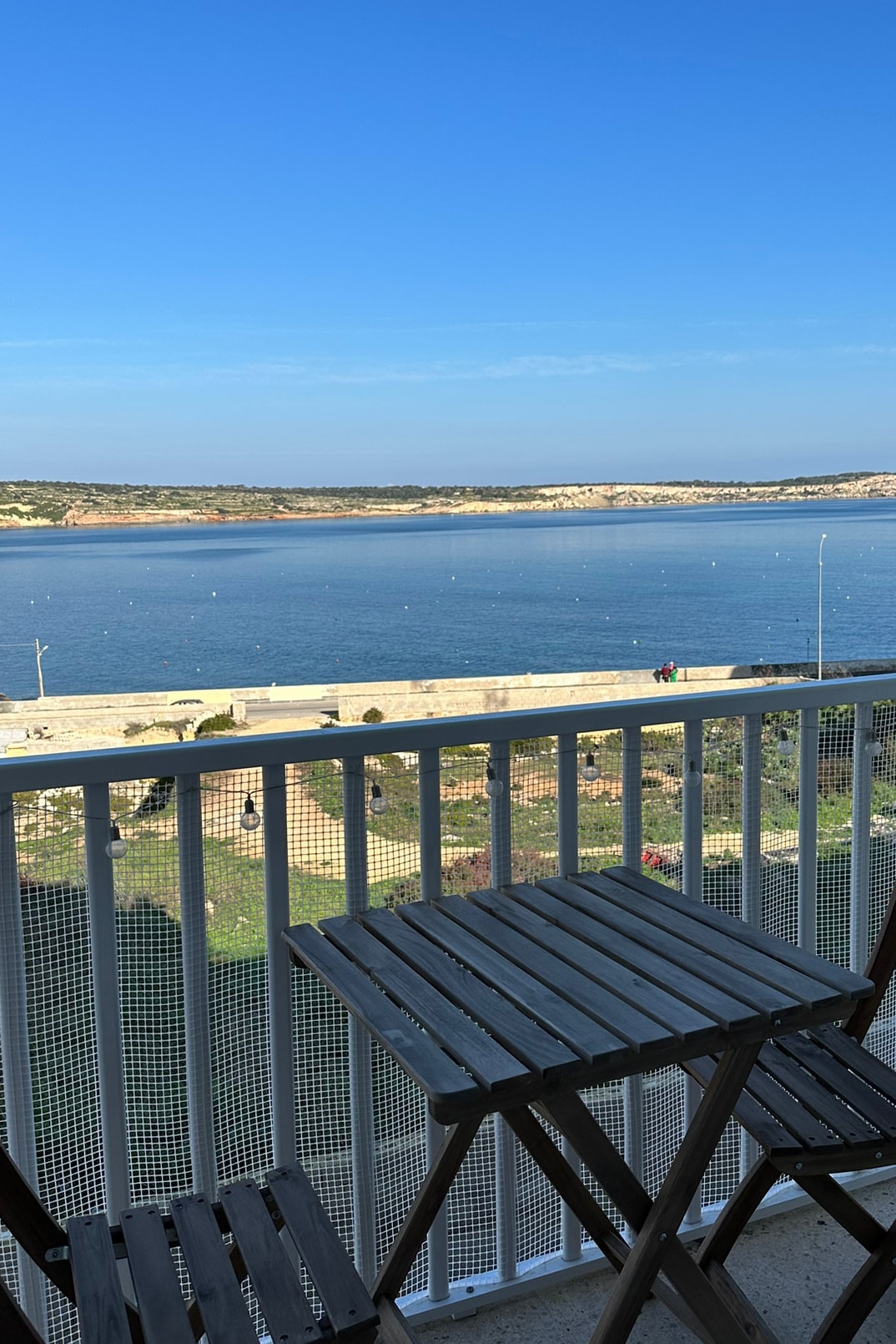 A balcony with two wooden chairs stands overlooking a calm sea, offering expansive views of the water and nearby shoreline. The sky is clear and blue, enhancing the serene atmosphere of the outdoor space.