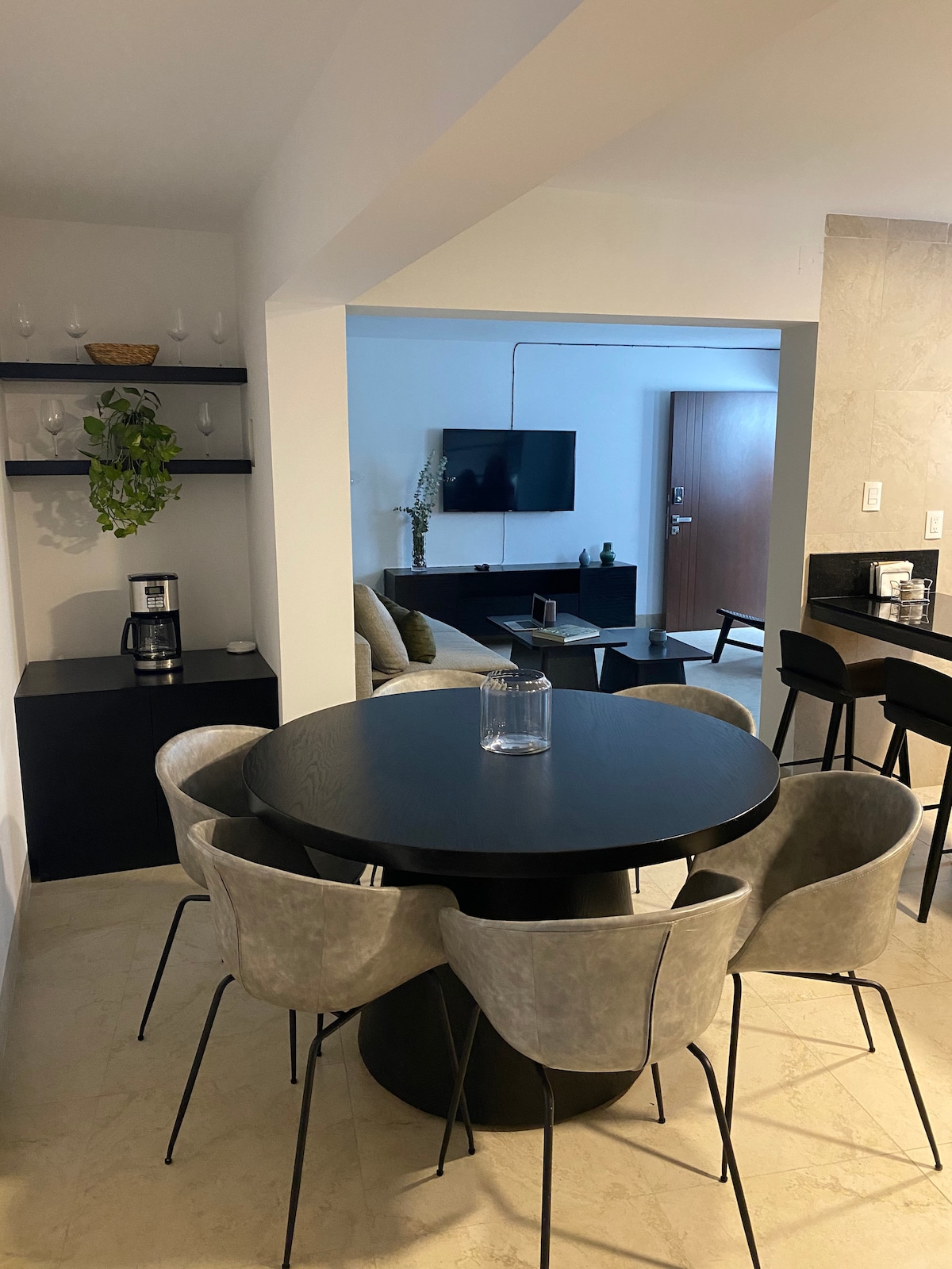 A modern dining area is featured with a round, dark table surrounded by four upholstered chairs. In the background, a cozy seating area is visible, accompanied by a television mounted on the wall. Decorative elements, including a plant and minimalist shelving, enhance the space.