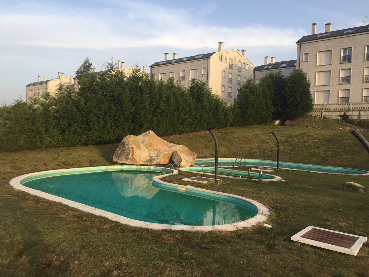 A large outdoor swimming pool is surrounded by manicured grass and natural stone features. The pool's unique shape is complemented bymultiple lounge areas, offering a serene setting. Nearby shrubbery and modern buildings create a tranquil backdrop.