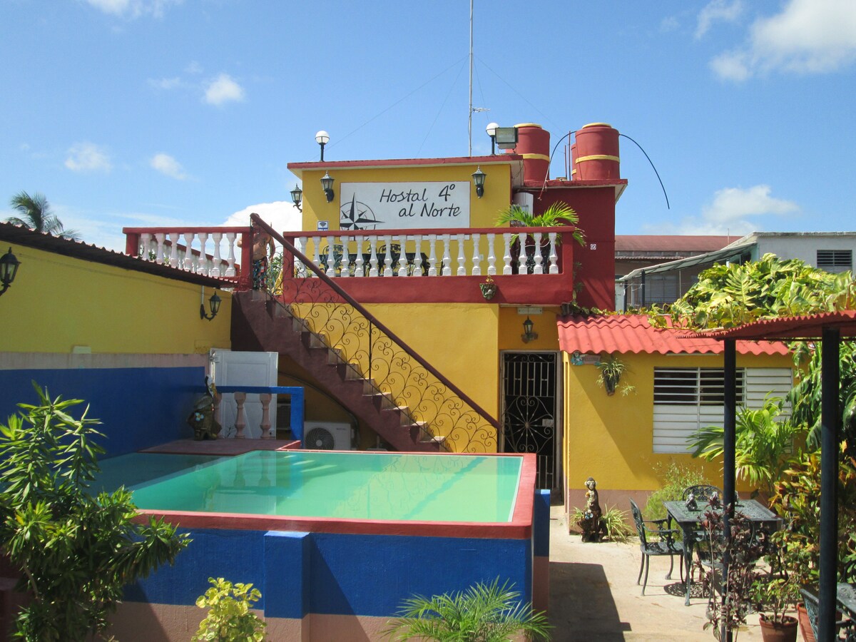 The exterior presents a colorful two-story building surrounded by vibrant landscaping. A refreshing pool is visible at the forefront, complemented by decorative plants. The entrance features a staircase leading to the second level, with inviting outdoor seating arrangements on the patio.