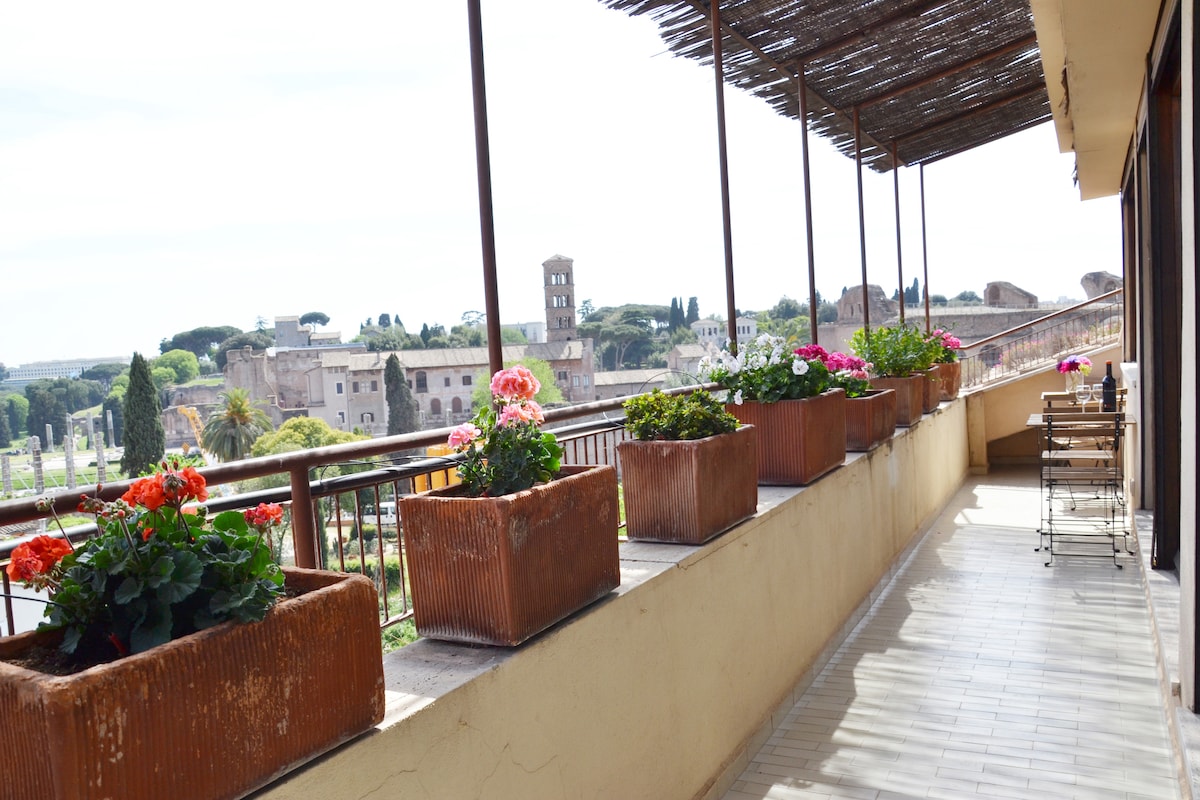 An outdoor terrace area is presented, featuring a railing lined with bloom-filled planters. The space is furnished with a small table and chairs, offering a direct view of historical landmarks. Natural light brightens the terrace, enhancing the vibrant floral accents.