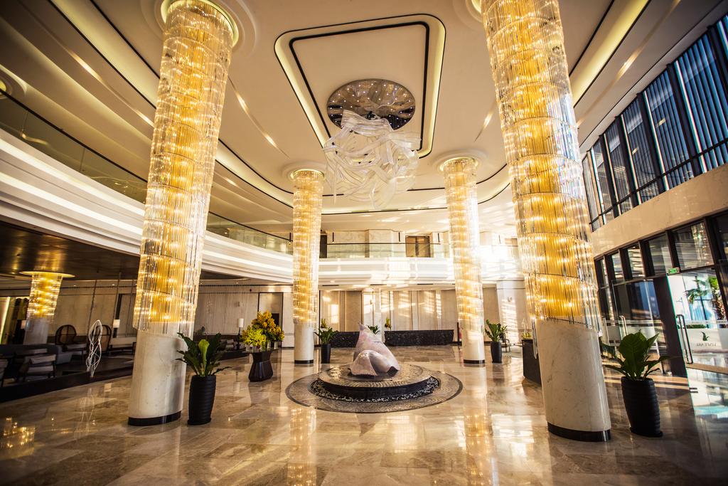 The spacious lobby features tall, illuminated pillars surrounded by elegant decor. A large decorative centerpiece is situated on the polished marble floor, complemented by lush greenery in the form of potted plants. Natural light streams in through expansive glass windows, creating an inviting atmosphere.