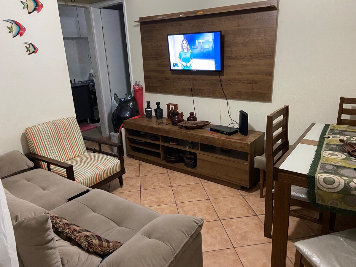 The living area features a comfortable sofa and striped chairs, aimed at promoting relaxation. A wooden TV unit holds a flat-screen television. A dining table with matching chairs occupies one side of the room, creating a cozy gathering space for meals.