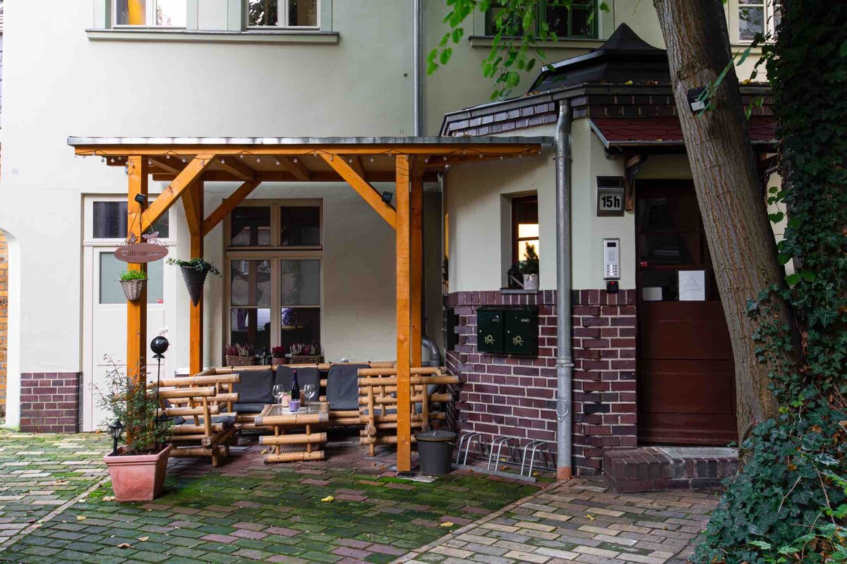 An inviting outdoor seating area is situated under a wooden awning, featuring a arrangement of wooden benches and a small table. Potted plants add greenery, while the surrounding stone pathway leads to the entrance of the building, framed by lush foliage.