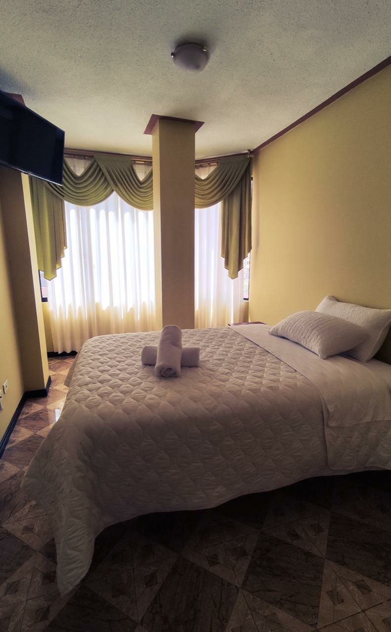 A cozy bedroom features a spacious bed dressed in neutral bedding. Large windows are adorned with soft, flowing drapes, allowing natural light to brighten the room. A television is mounted on the wall opposite the bed, enhancing the functionality of the space.