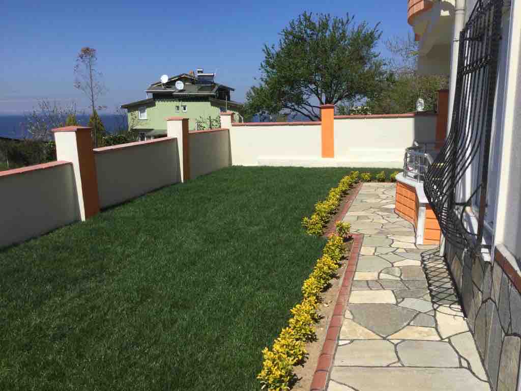 A well-maintained outdoor space is presented, featuring a manicured lawn bordered by neatly arranged stone pathways. Colorful shrubs line the edge of the garden, complementing the vibrant greenery. A glimpse of neighboring houses can be seen in the background under a clear blue sky.