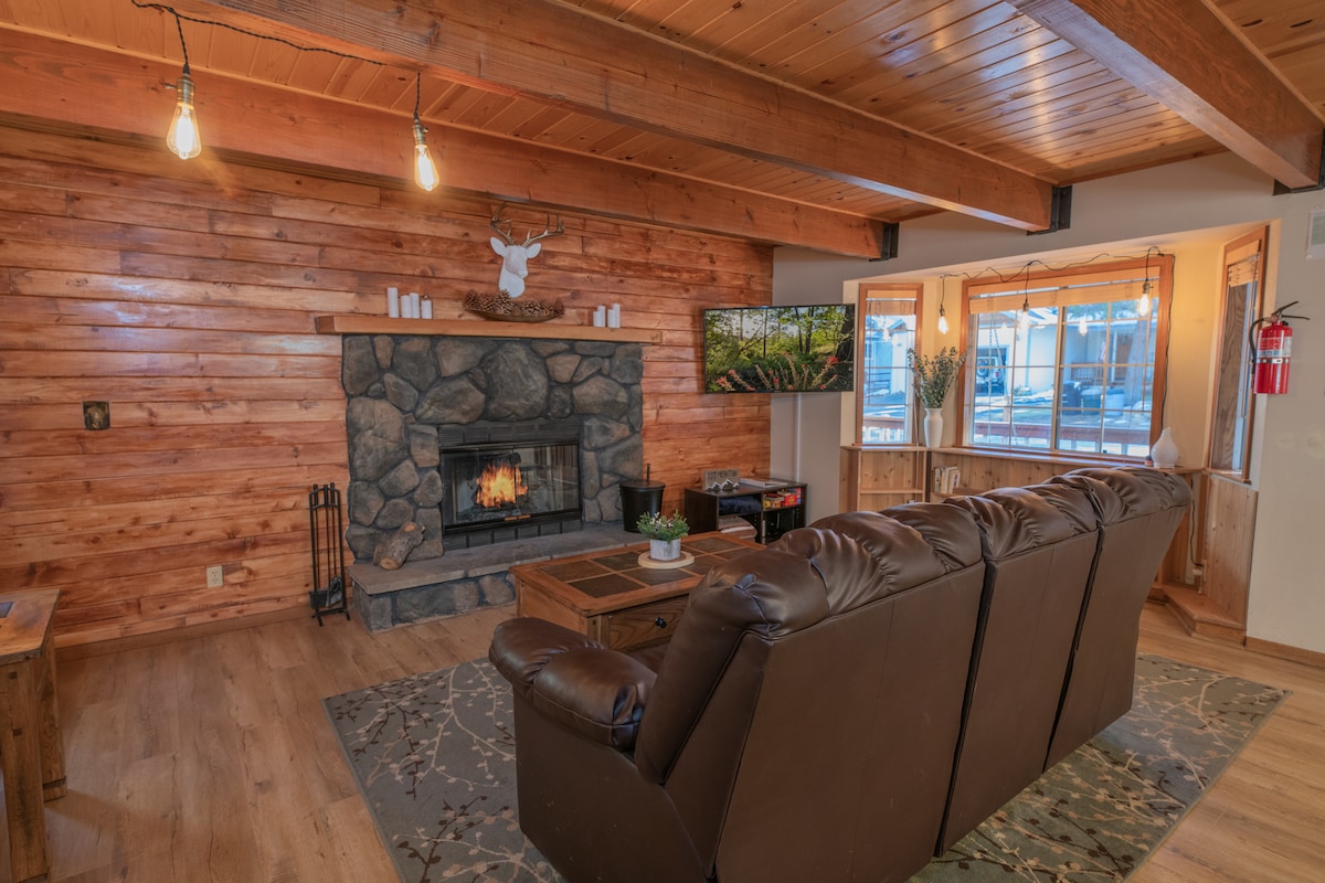 The living room features a stone fireplace, providing warmth and a natural focal point. Large windows bring in plenty of light, while wooden beams and walls create a rustic charm. A comfortable brown couch faces the fireplace, inviting relaxation.