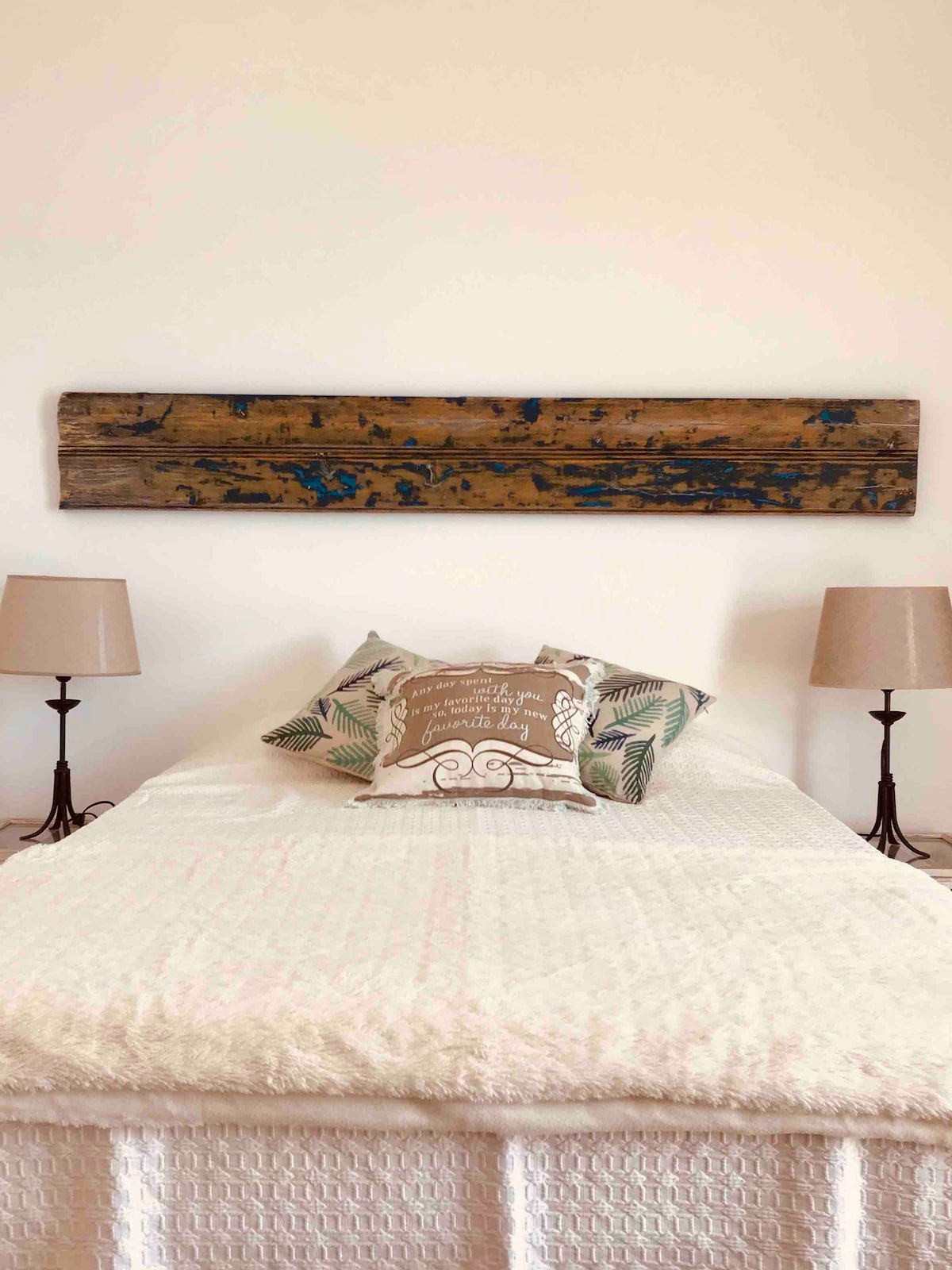 A spacious bed is positioned between two bedside lamps, providing soft illumination. A decorative wooden plank hangs above the bed, contrasting with the light-colored bedding. Soft throw pillows add a touch of comfort to the inviting space.