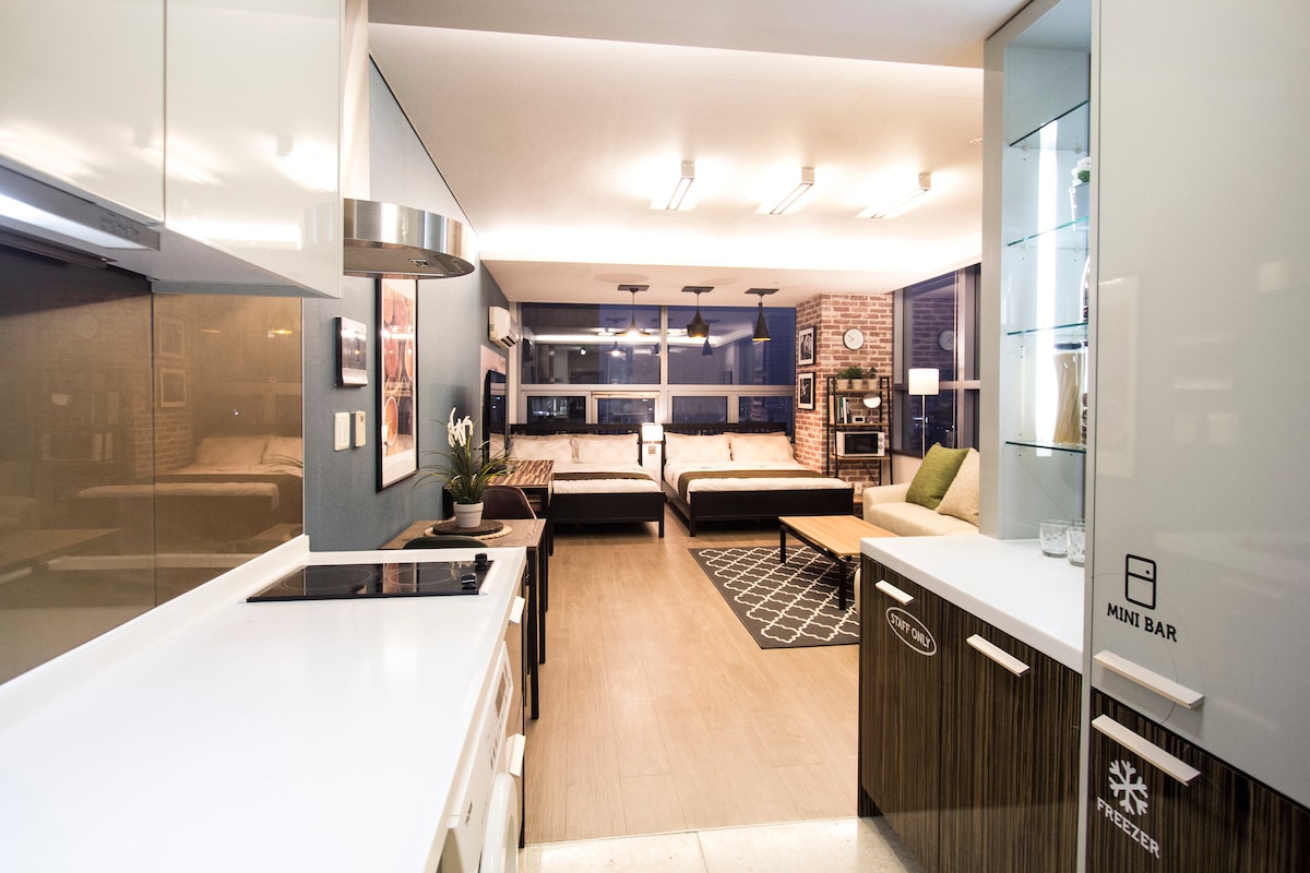 A modern kitchen space features sleek cabinetry and a built-in mini bar. The open layout connects to a living area with two spacious beds, offering natural light through large windows. A textured rug adds a cozy aspect, complementing the contemporary design aesthetic.