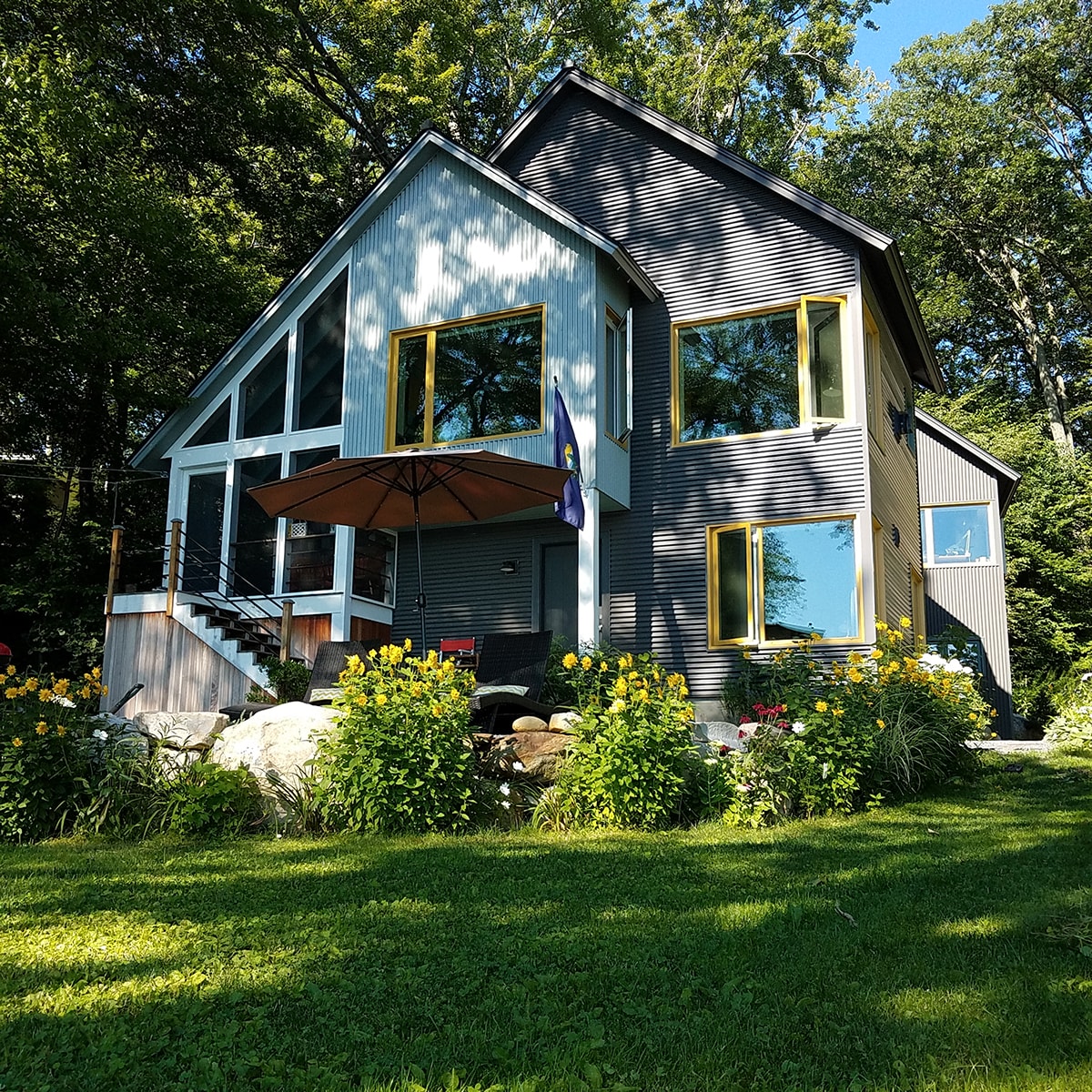 A modern house features a combination of gray siding and large windows, surrounded by lush greenery and colorful flowers. A welcoming entryway with a staircase is visible, and an umbrella shades a seating area on the porch, creating an inviting outdoor space.