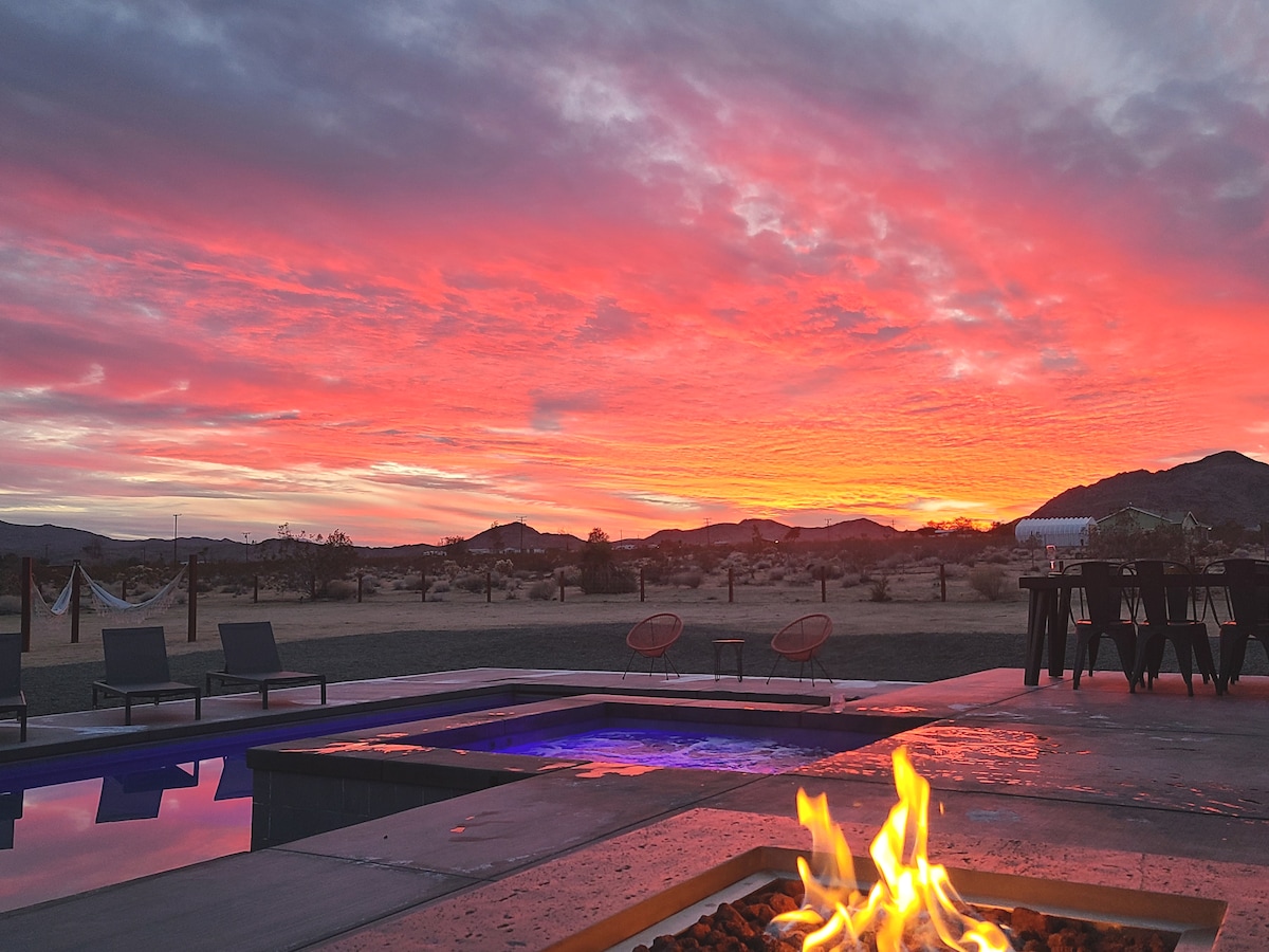 A vibrant sunset paints the sky in hues of orange and pink, viewed from the outdoor area. A warm fire pit crackles in the foreground, with the in-ground pool and hot tub reflecting the colorful sky. Lounge chairs and hammocks are visible in the expansive desert landscape.