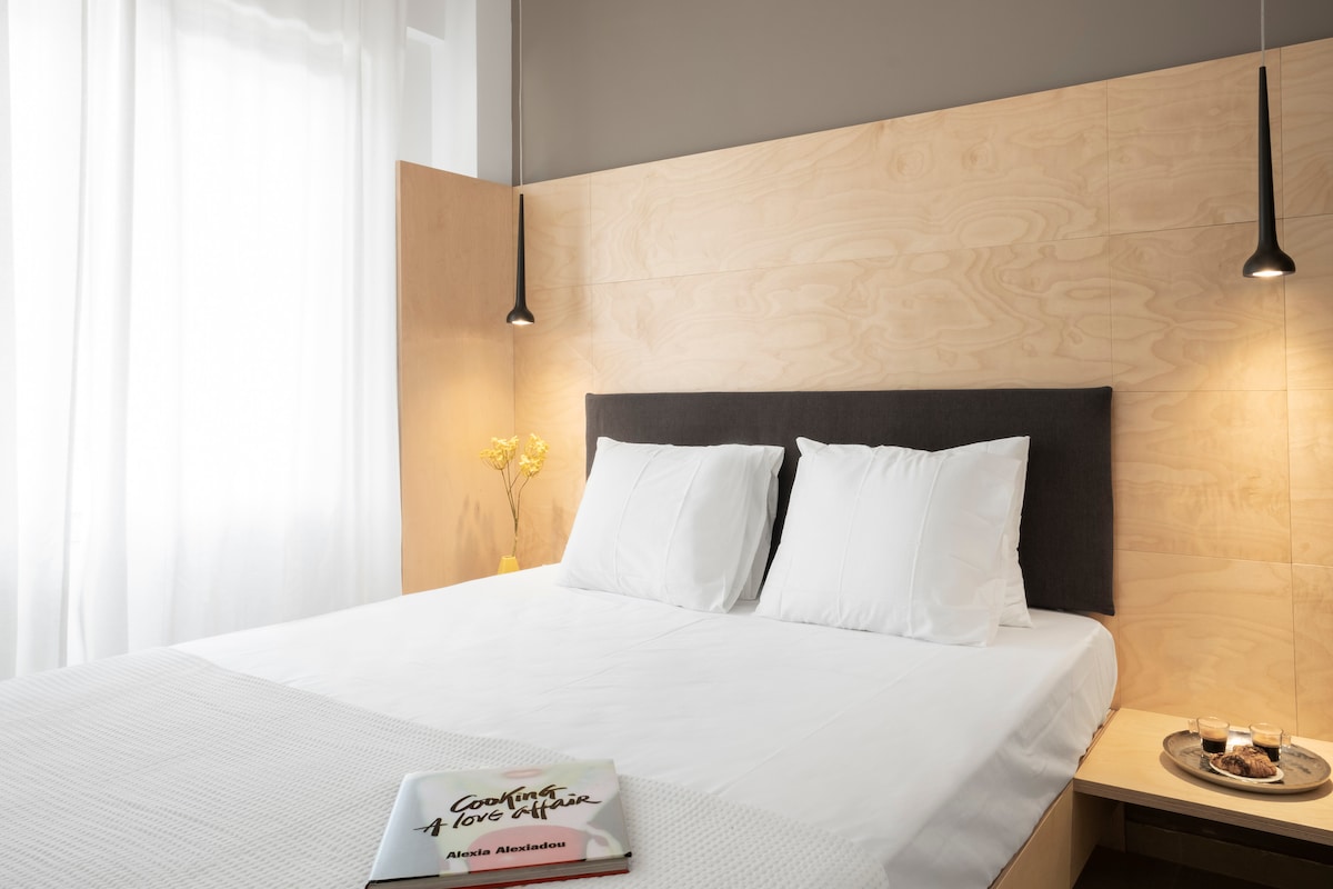 A minimalistic bedroom displays a spacious bed with crisp white linens and two plush pillows. Soft, natural light filters through sheer curtains, illuminating the light wooden panel behind the bed. A small bedside table holds a book and a decorative item, creating a serene and uncluttered atmosphere.