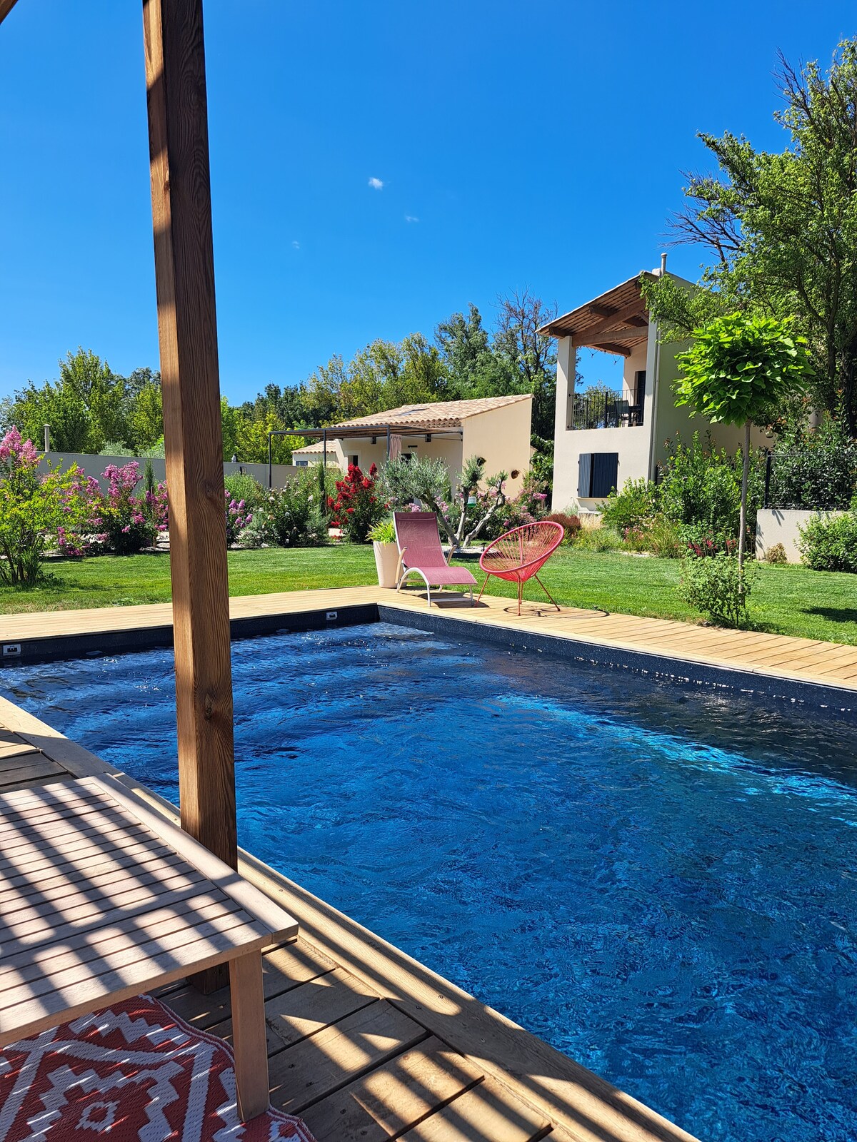 A serene outdoor space features a salt-water pool surrounded by lush greenery. The wooden deck is complemented by a stylish lounge chair and a pink chair, inviting relaxation under clear blue skies.